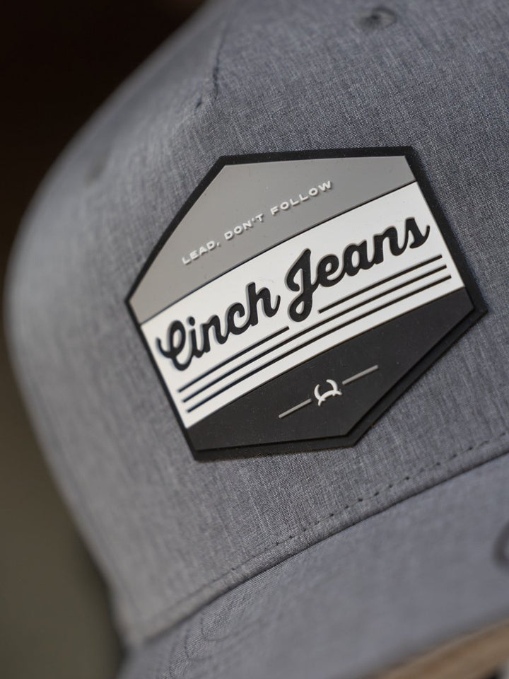 Close-up of the Cinch Lead Cap by Savannah Sevens western life{&}style, a gray trucker hat with a hexagonal patch reading "Lead. Dont Follow" and "Cinch Jeans," featuring black/white stripes, emblem, and adjustable snap back.