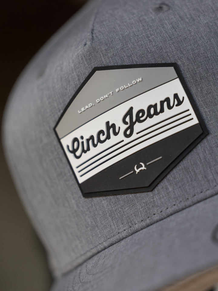 Close-up of the Cinch Lead Cap by Savannah Sevens western life{&}style, a gray trucker hat with a hexagonal patch reading "Lead. Dont Follow" and "Cinch Jeans," featuring black/white stripes, emblem, and adjustable snap back.