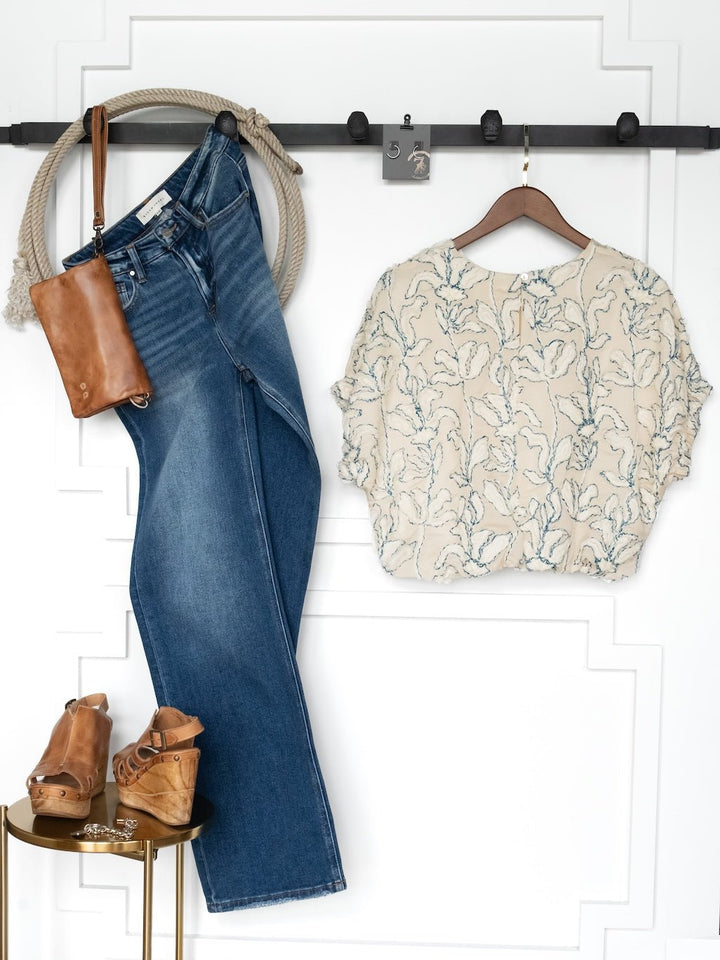 The Clare Textured Floral Bubble Hem Top by Savannah Sevens western life{&}style hangs with blue jeans, a brown leather crossbody bag, a tan rope belt, and tan wedge sandals against a white paneled wall.