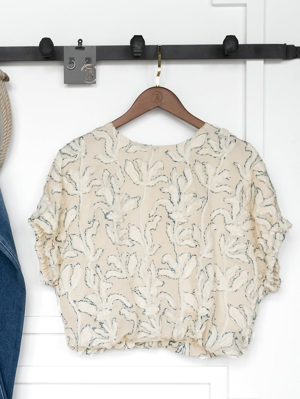 The Clare Textured Floral Bubble Hem Top by Savannah Sevens western life{&}style, a cream relaxed-fit blouse with a floral pattern, hangs on a wooden hanger against a white wall beneath a black metal hook rack.