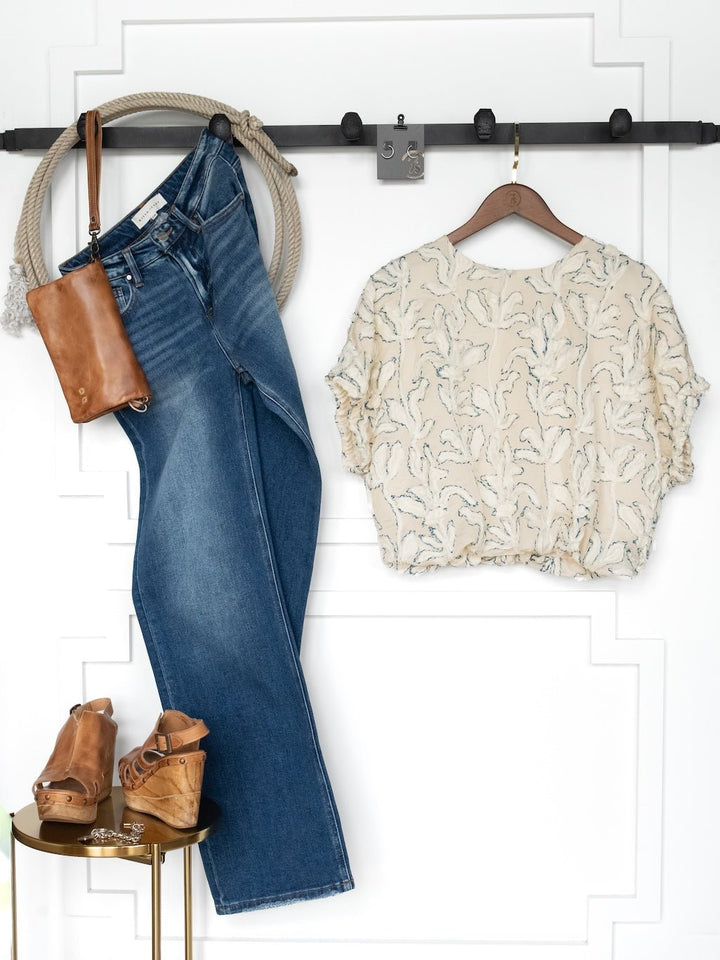The Clare Textured Floral Bubble Hem Top by Savannah Sevens western life{&}style hangs with blue jeans, tan wedge sandals, a brown leather crossbody bag, and a rope accessory against a white paneled wall.