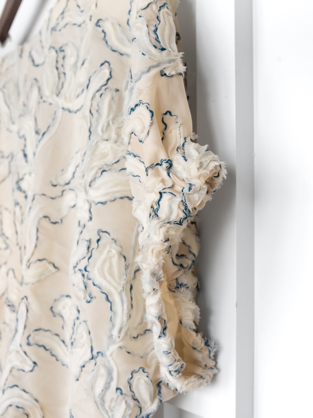 The Clare Textured Floral Bubble Hem Top by Savannah Sevens western life{&}style features cream-colored, semi-sheer fabric with frayed floral patterns and blue embroidered outlines, shown hanging on a white surface.