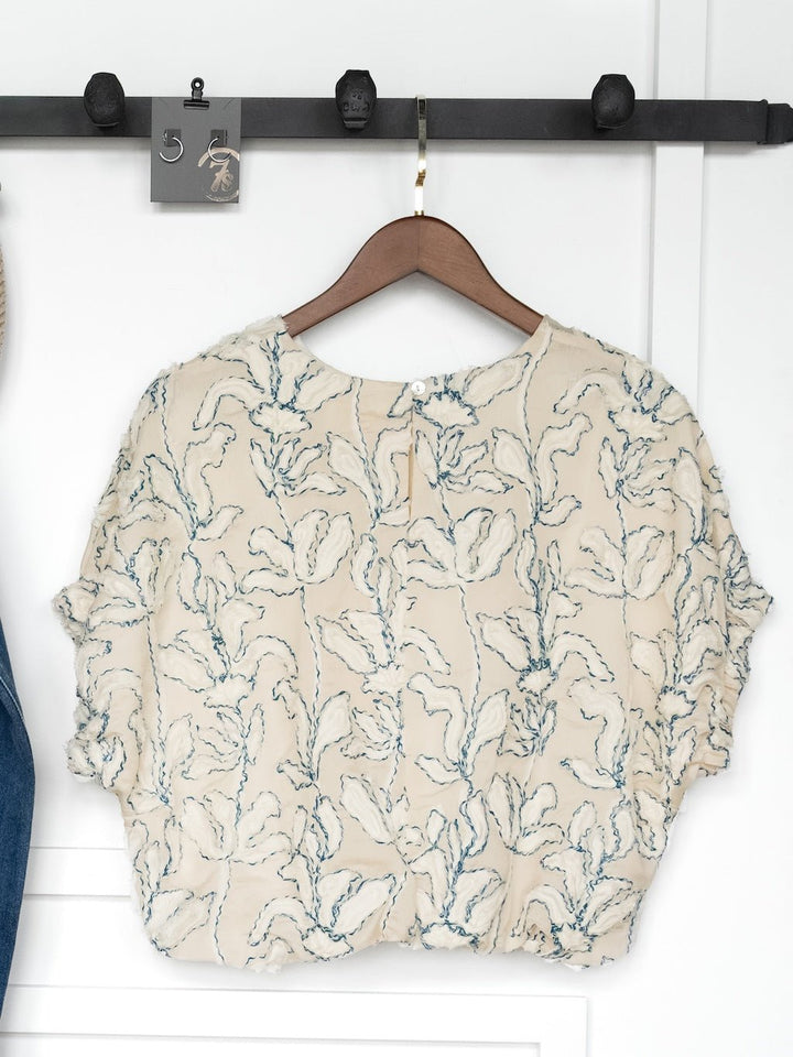 The Clare Textured Floral Bubble Hem Top by Savannah Sevens western life{&}style, in cream with blue floral embroidery, hangs on a wooden hanger from a black wall rail. A tag labeled “5” is clipped above the blouse.