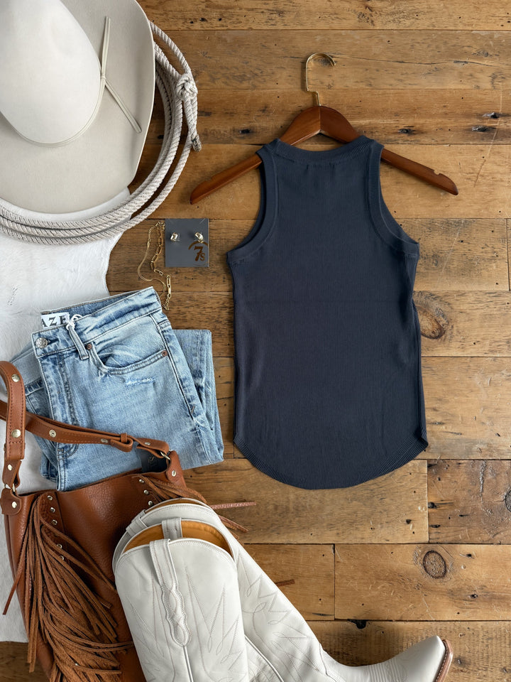 Flat lay of the Clermont Charcoal Ribbed Tank by Savannah Sevens western life{&}style on a wooden surface, styled with light denim jeans, a white cowboy hat, rope, white cowboy boots, a brown fringe bag, and gold jewelry arranged beside it.