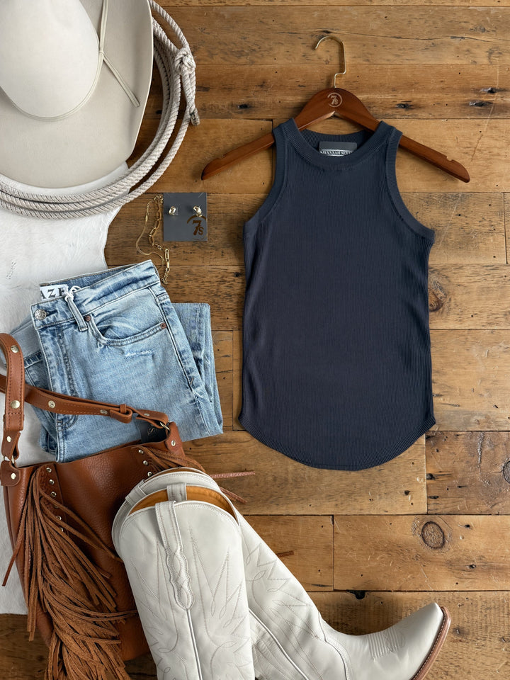 The Clermont Charcoal Ribbed Tank by Savannah Sevens western life{&}style is displayed on a hanger alongside light wash jeans, a brown fringe bag, white cowboy boots, a hat, rope, and an earring set—all arranged in a Western-inspired flat lay on a wooden floor.