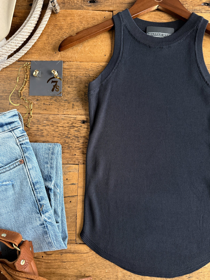 The Clermont Charcoal Ribbed Tank by Savannah Sevens western life{&}style is displayed on a hanger alongside light blue jeans, gold jewelry, a brown belt, and a beige bag arranged on a wooden surface.