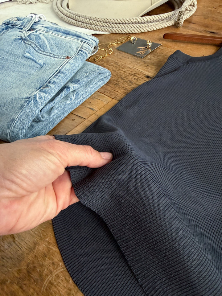 A hand holds the Clermont Charcoal Ribbed Tank by Savannah Sevens western life{&}style on a wooden table, with folded jeans, a chain necklace, square earrings, and a beige rope belt in the background.