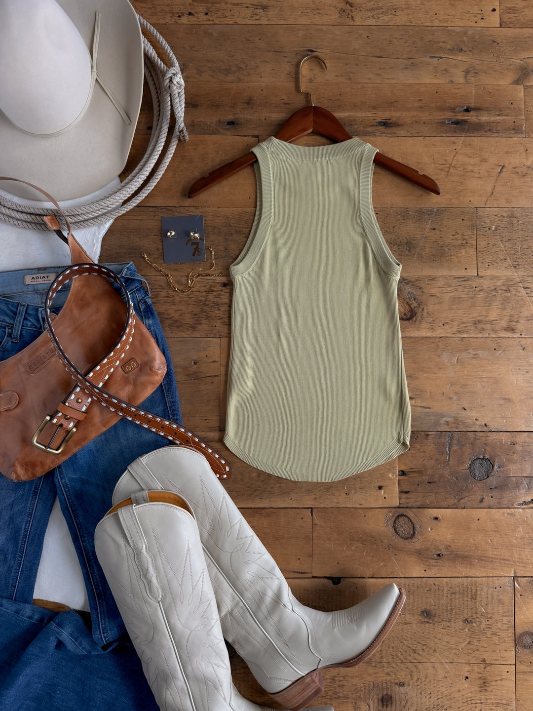 Flat lay of a western-inspired outfit featuring cream cowboy boots, blue jeans, the Clermont Sage Ribbed Tank from Savannah Sevens western life{&}style on a hanger, a brown leather purse, white cowboy hat, and gold jewelry displayed on a rustic wooden floor.