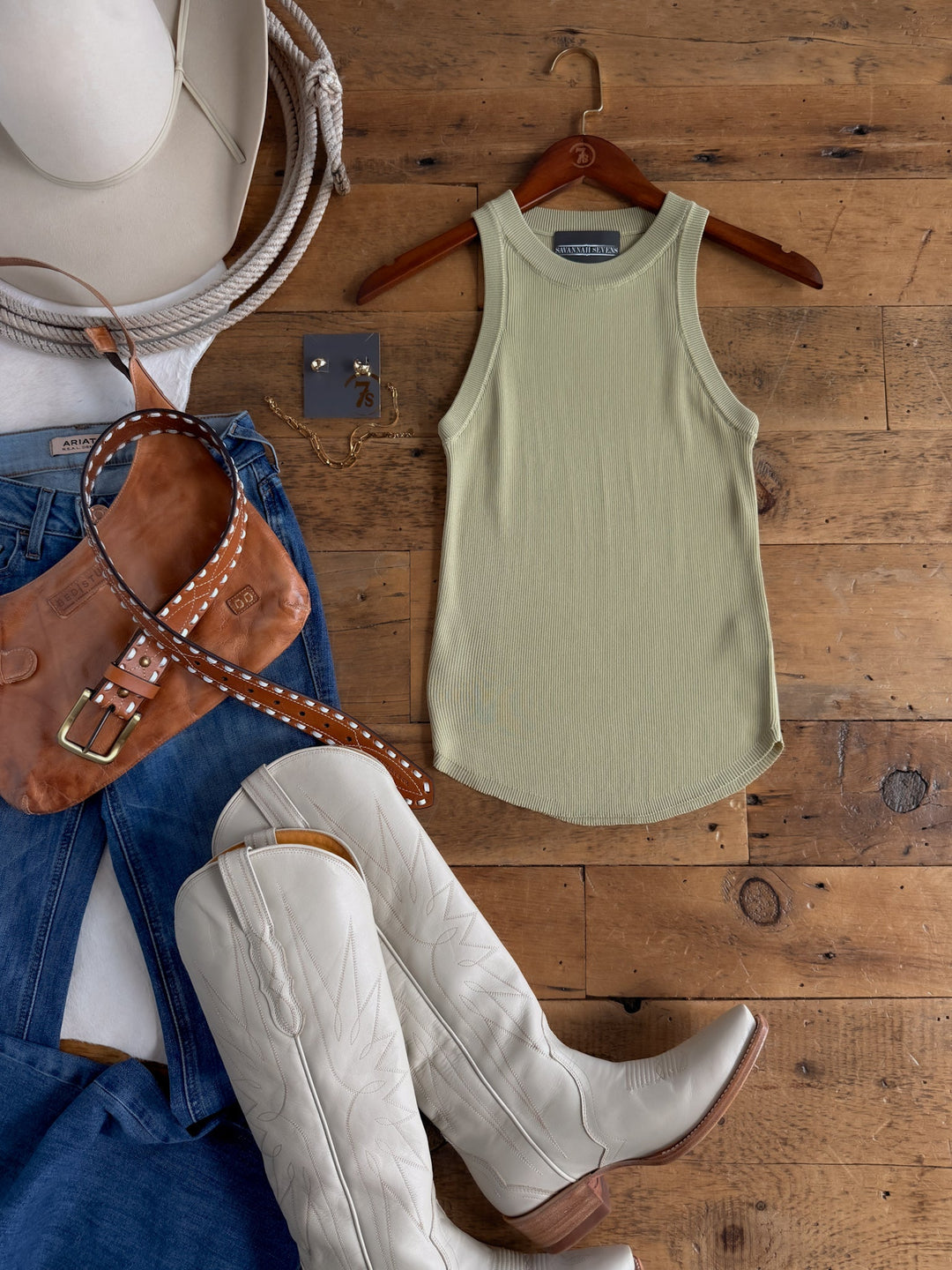 Flat lay of a Western-style outfit featuring the Savannah Sevens western life{&}style Clermont Sage Ribbed Tank, a cream cowboy hat, white cowboy boots, blue jeans, a brown leather purse with a studded strap, and gold earrings arranged on a wooden floor.