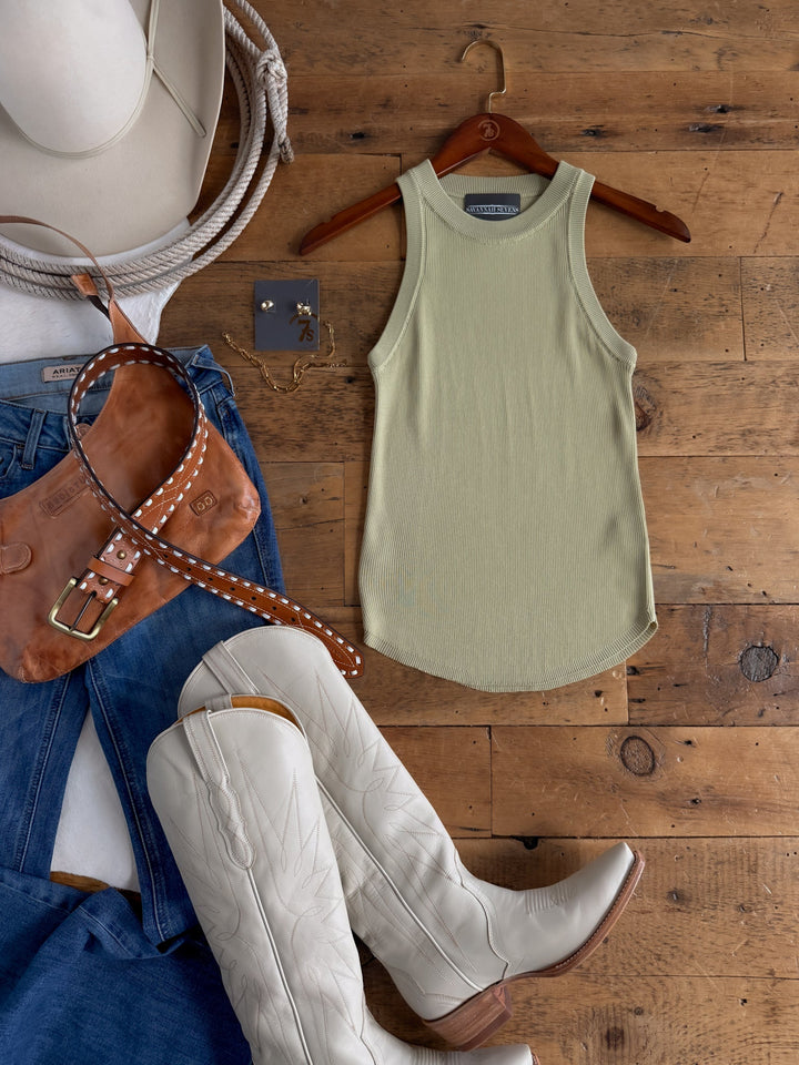 Flat lay of a Western-style outfit featuring the Savannah Sevens western life{&}style Clermont Sage Ribbed Tank, a cream cowboy hat, white cowboy boots, blue jeans, a brown leather purse with a studded strap, and gold earrings arranged on a wooden floor.