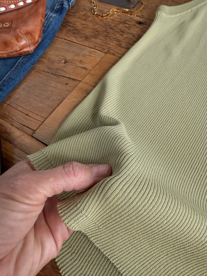 A hand gently pinches the ribbed fabric of the Clermont Sage Ribbed Tank by Savannah Sevens western life{&}style, displayed on a wooden surface alongside a brown leather bag, blue jeans, and a gold chain necklace.