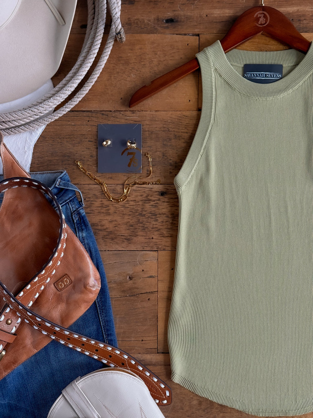 A flat lay of the Clermont Sage Ribbed Tank by Savannah Sevens western life{&}style on a wooden surface, styled with jeans, a brown leather bag, a white hat, gold jewelry, and a belt adorned with silver studs.