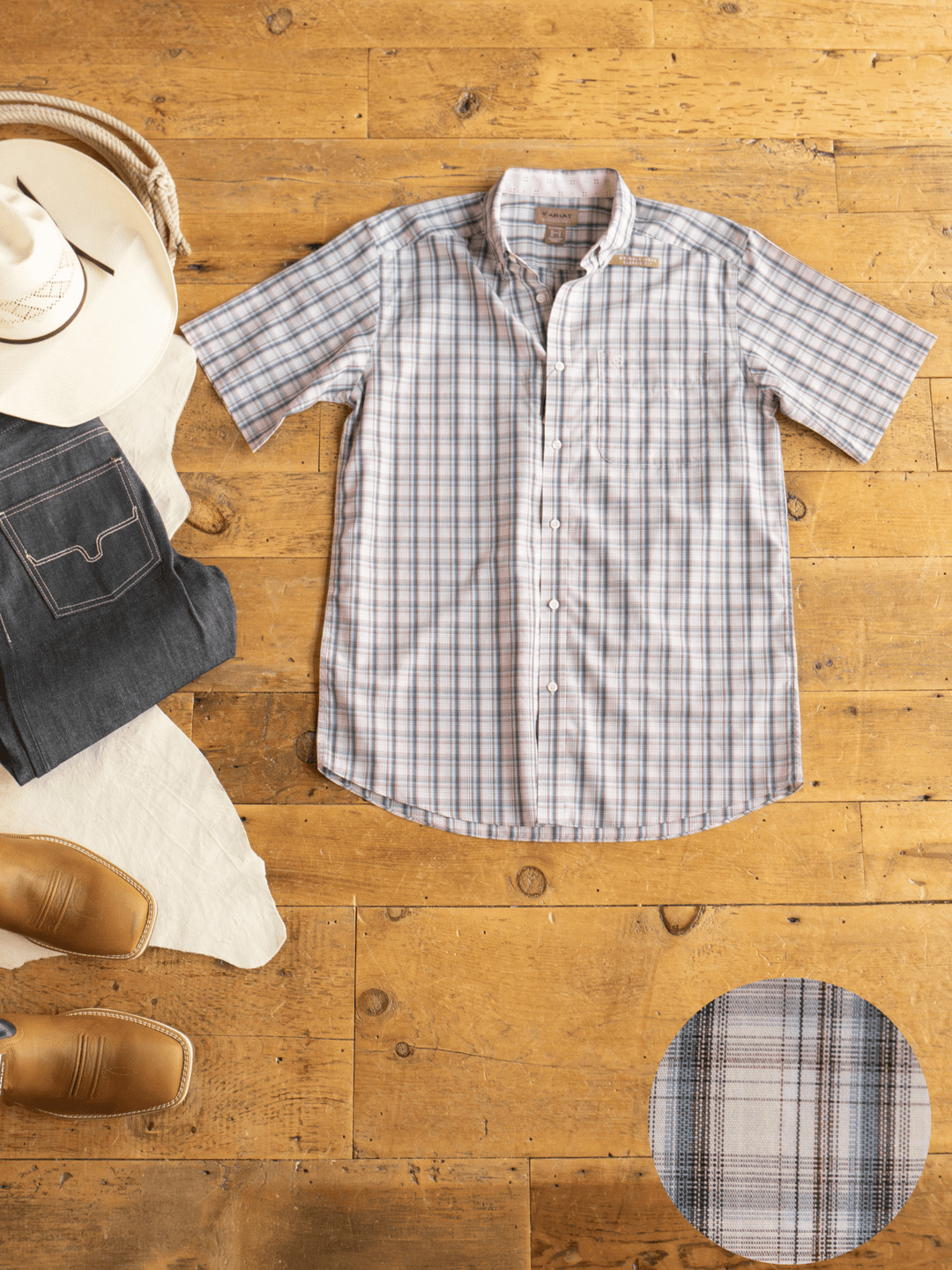 The Savannah Sevens western life{&}style Clifton Ariat Men's Plaid Button Up is laid flat on a wooden floor with jeans, tan cowboy boots, and a white cowboy hat. An inset highlights the shirt’s plaid fabric pattern.