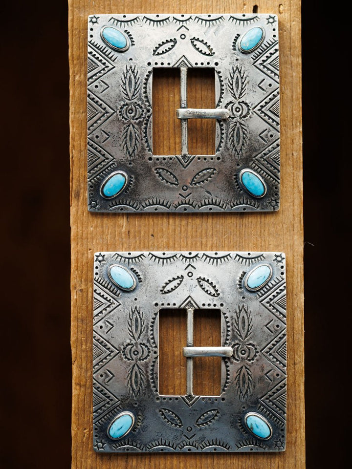 Two ornate, square Coalinga Buckles by Savannah Sevens western life{&}style, featuring engraved sterling silver and turquoise stones at each corner, are displayed vertically on a wooden plank.