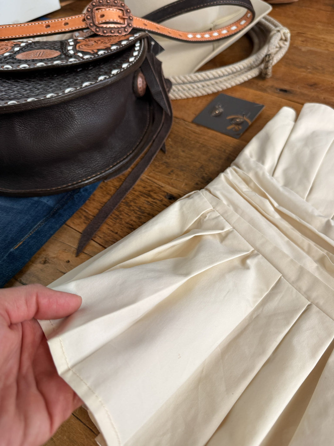 A hand holds the Cohutta Cream Strapless Top from Savannah Sevens western life{&}style. Nearby are a brown leather bag with studs, a beige purse, small earrings, and a pair of denim jeans on a wooden surface.
