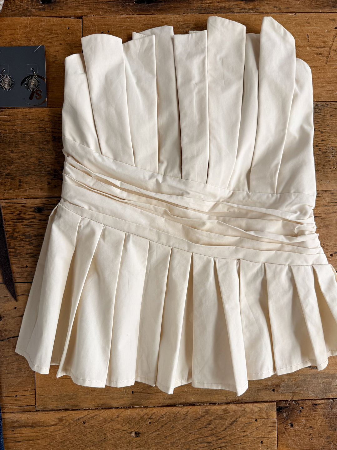 The Cohutta Cream Strapless Top by Savannah Sevens western life{&}style, featuring a pleat design with a fitted bodice and flared hem, is displayed on a wooden surface. A pair of earrings appears in the upper left corner.