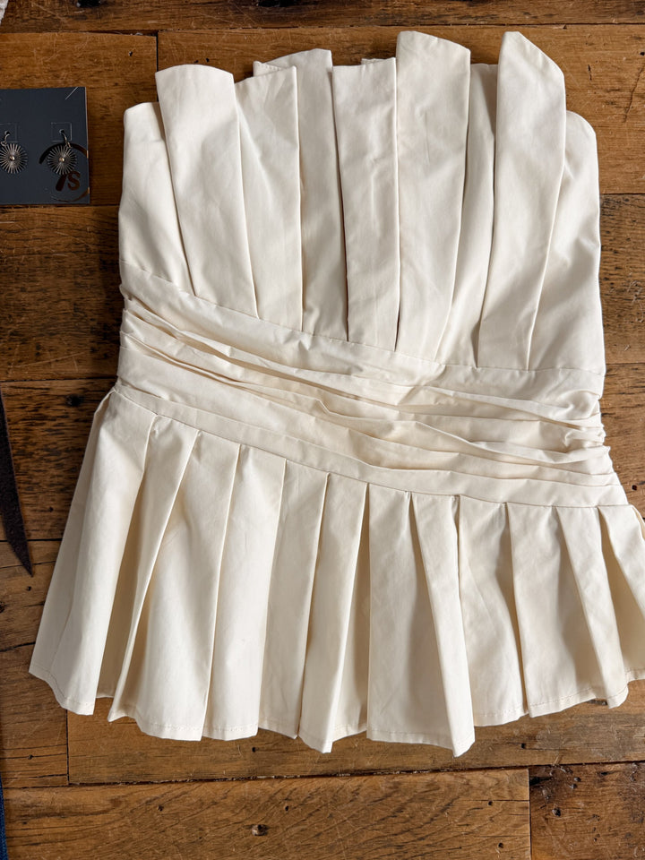 The Cohutta Cream Strapless Top by Savannah Sevens western life{&}style, featuring a pleat design with a fitted bodice and flared hem, is displayed on a wooden surface. A pair of earrings appears in the upper left corner.
