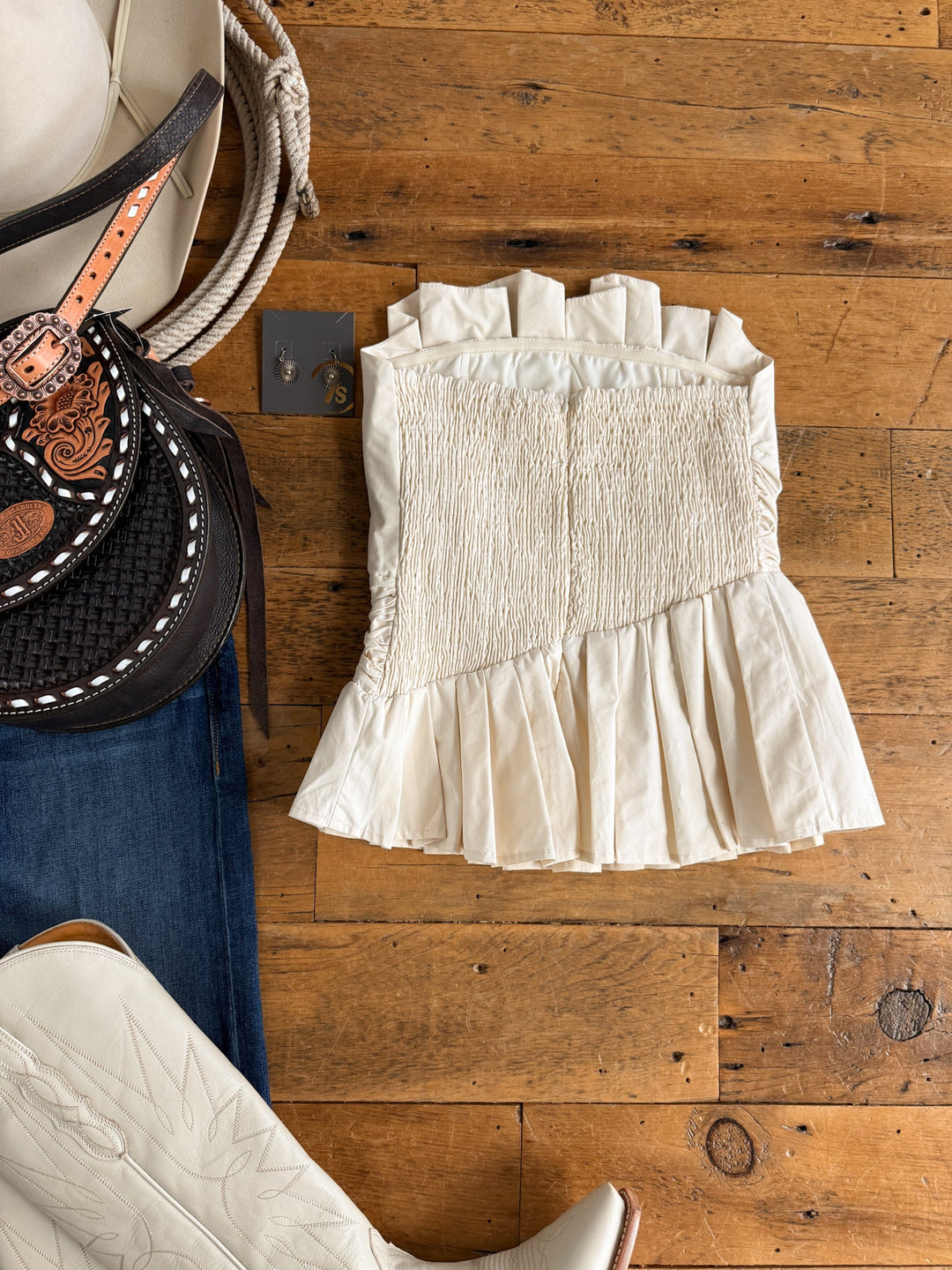 Cohutta Cream Strapless Top by Savannah Sevens western life{&}style, paired with white cowboy boots, dark jeans, a Western-style hat, rope, a tooled leather bag, and earrings arranged on a wooden floor.