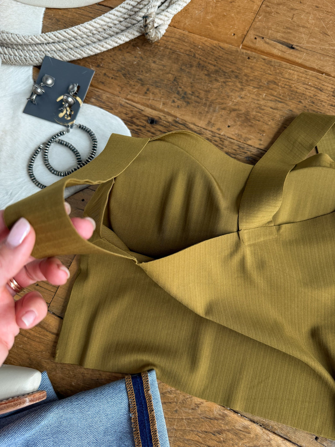 A hand holds the strap of the Coleta Chartreuse Olive Brami by Savannah Sevens western life{&}style, displayed on a wooden surface alongside jewelry, denim fabric, rope, and a small white pouch.