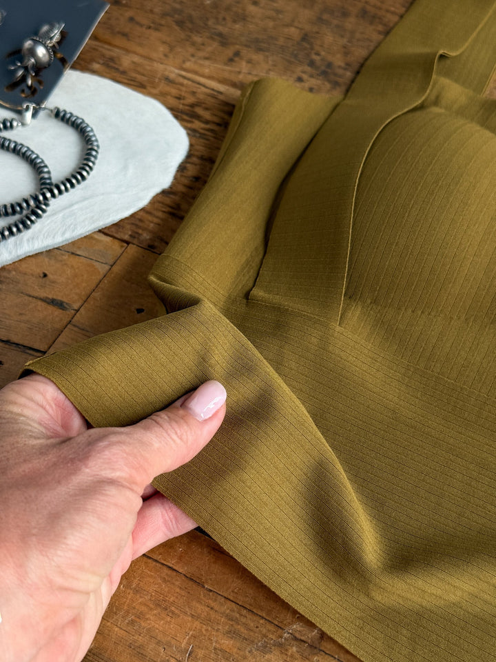 A hand holds the stretch fabric of the Coleta Chartreuse Olive Brami by Savannah Sevens western life{&}style, showing its ribbed texture on a wooden surface. A silver necklace and part of a device are visible nearby.