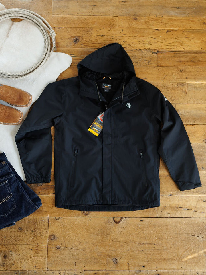 A Collbran Ariat men's black jacket by Savannah Sevens western life{&}style with tags is laid flat on a wooden floor beside a white cowhide rug, tan sandals, and folded dark blue jeans.