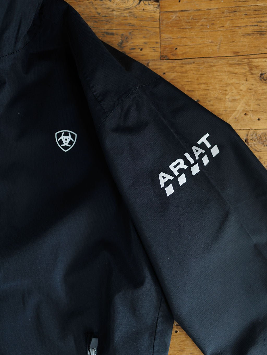 A close-up of the Collbran Ariat Men's Black Jacket by Savannah Sevens western life{&}style shows the ARIAT logo, a white checkered sleeve design, and a small shield emblem on the chest, displayed on a wooden surface.