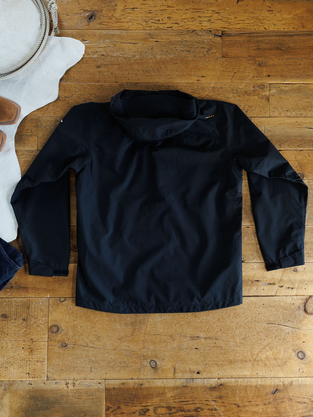 A Collbran Ariat Men's Black Jacket by Savannah Sevens western life{&}style, with a hood and long sleeves, is laid flat on a wooden floor with part of a white rug and brown sandals visible in the corner.