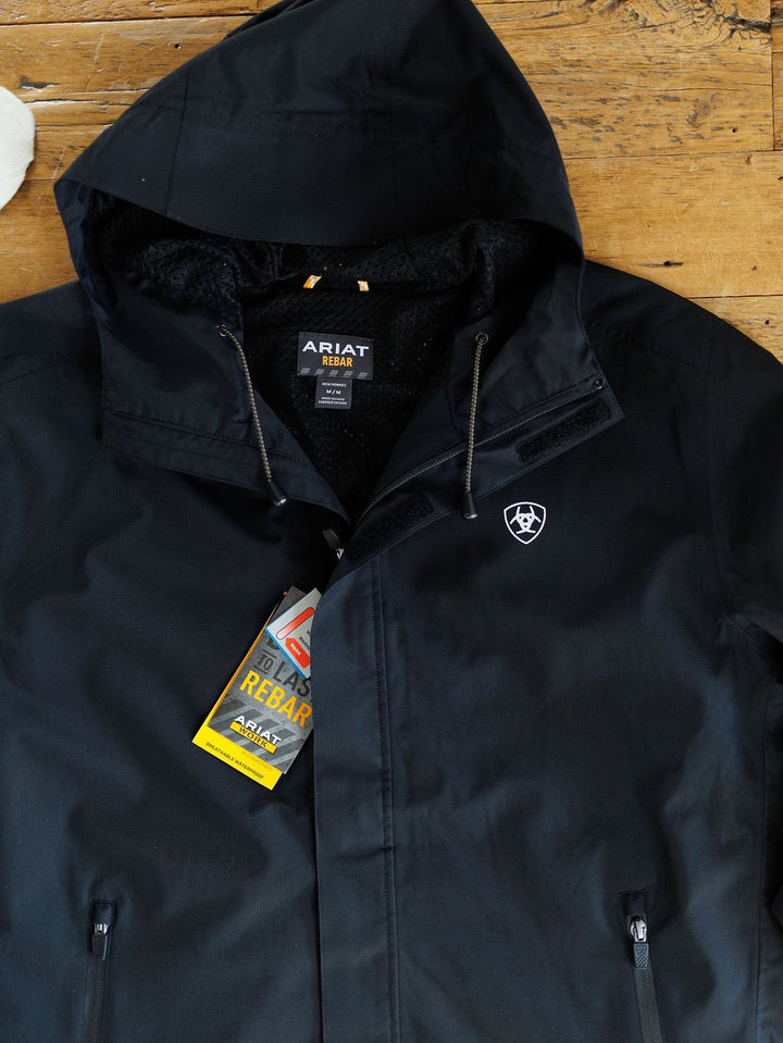 A Collbran Ariat Men's Black Jacket by Savannah Sevens western life{&}style is laid flat on a wooden surface, displaying its chest logo, zippers, and tags. This jacket is windproof, waterproof, stylish, and durable.