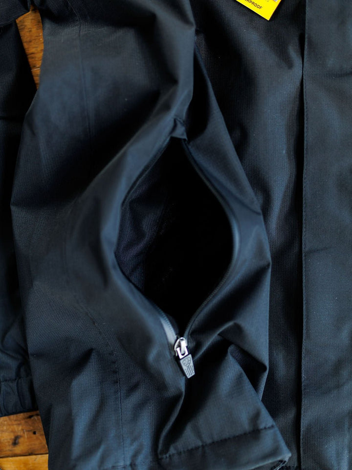 Close-up of the Collbran Ariat Men's Black Jacket by Savannah Sevens western life{&}style, showing a partially unzipped pocket with visible inner lining, zipper pull, and a hint of a yellow tag near the top right corner.