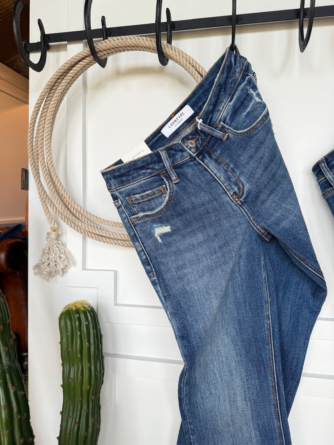 The Collins Premier Rise Straight Leg Jean by Savannah Sevens western life{&}style hangs by a belt loop from a rope hook, with a green cactus and another pair of straight leg jeans nearby.