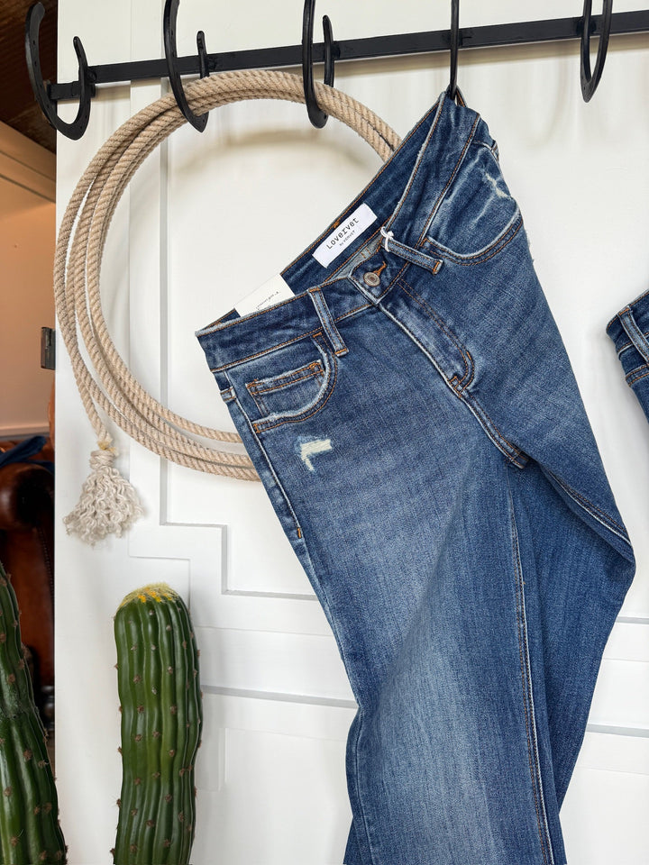 The Collins Premier Rise Straight Leg Jean by Savannah Sevens western life{&}style hangs by a belt loop from a rope hook, with a green cactus and another pair of straight leg jeans nearby.