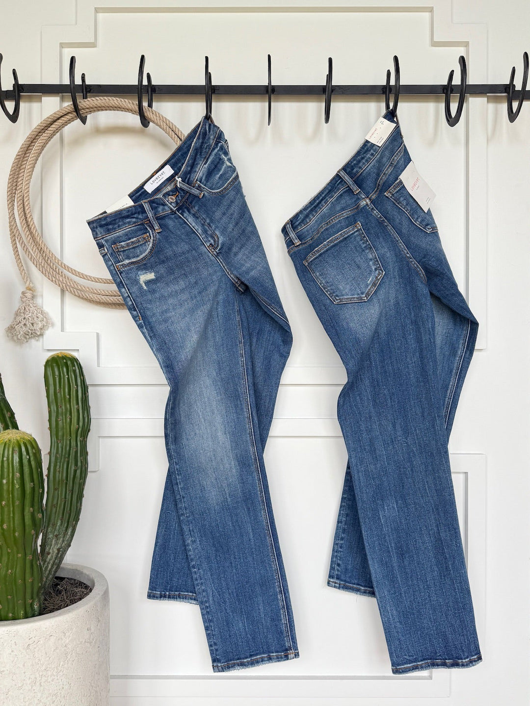 Two pairs of The Collins Premier Rise Straight Leg Jean by Savannah Sevens western life{&}style in a medium dark wash hang from black hooks on a white wall, with a coiled rope and potted cactus below, all bathed in natural light.