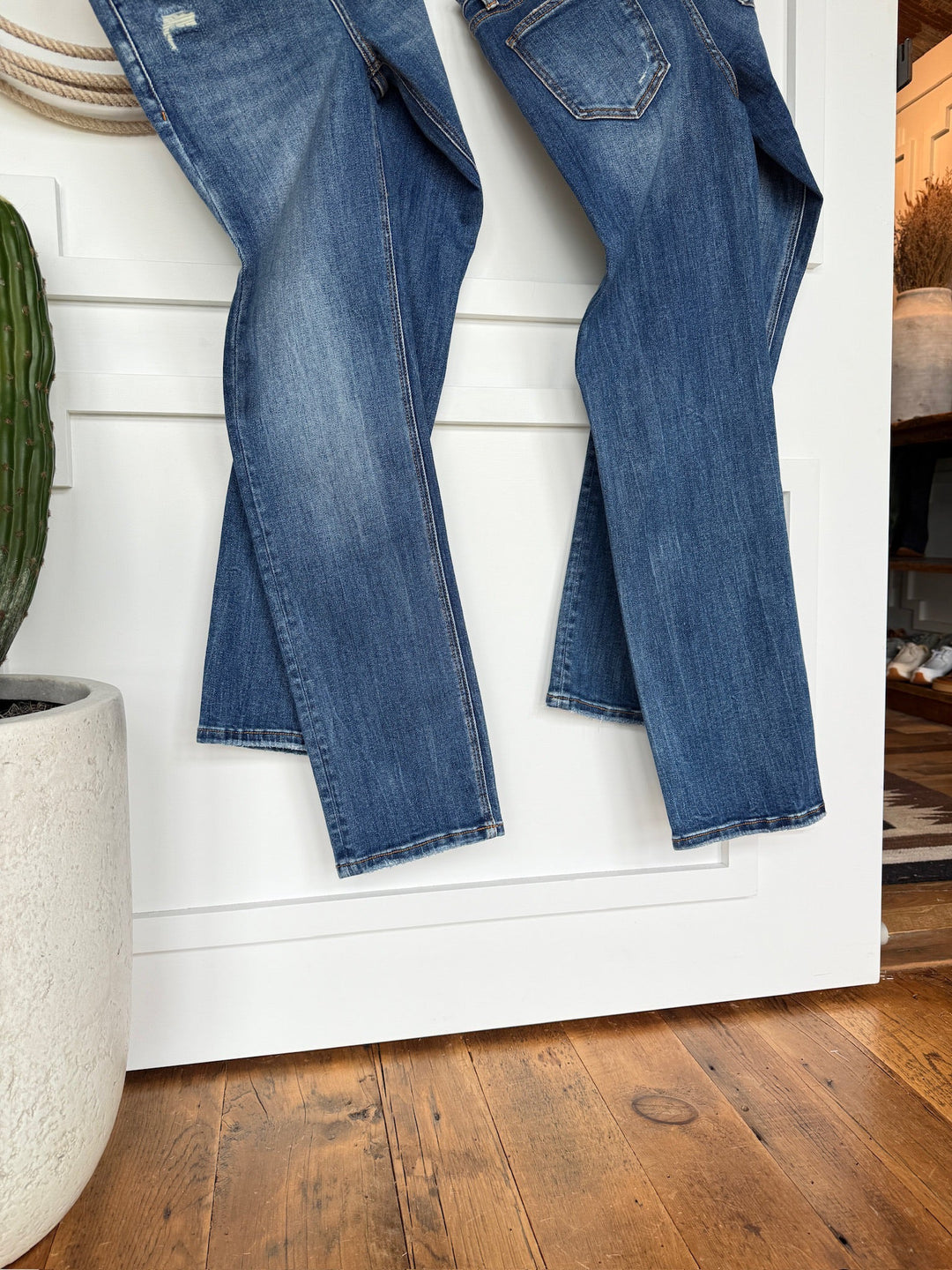 The Collins Premier Rise Straight Leg Jean by Savannah Sevens western life{&}style in a medium dark wash hangs on a white door, with a large potted cactus beside it and a wooden floor underneath.