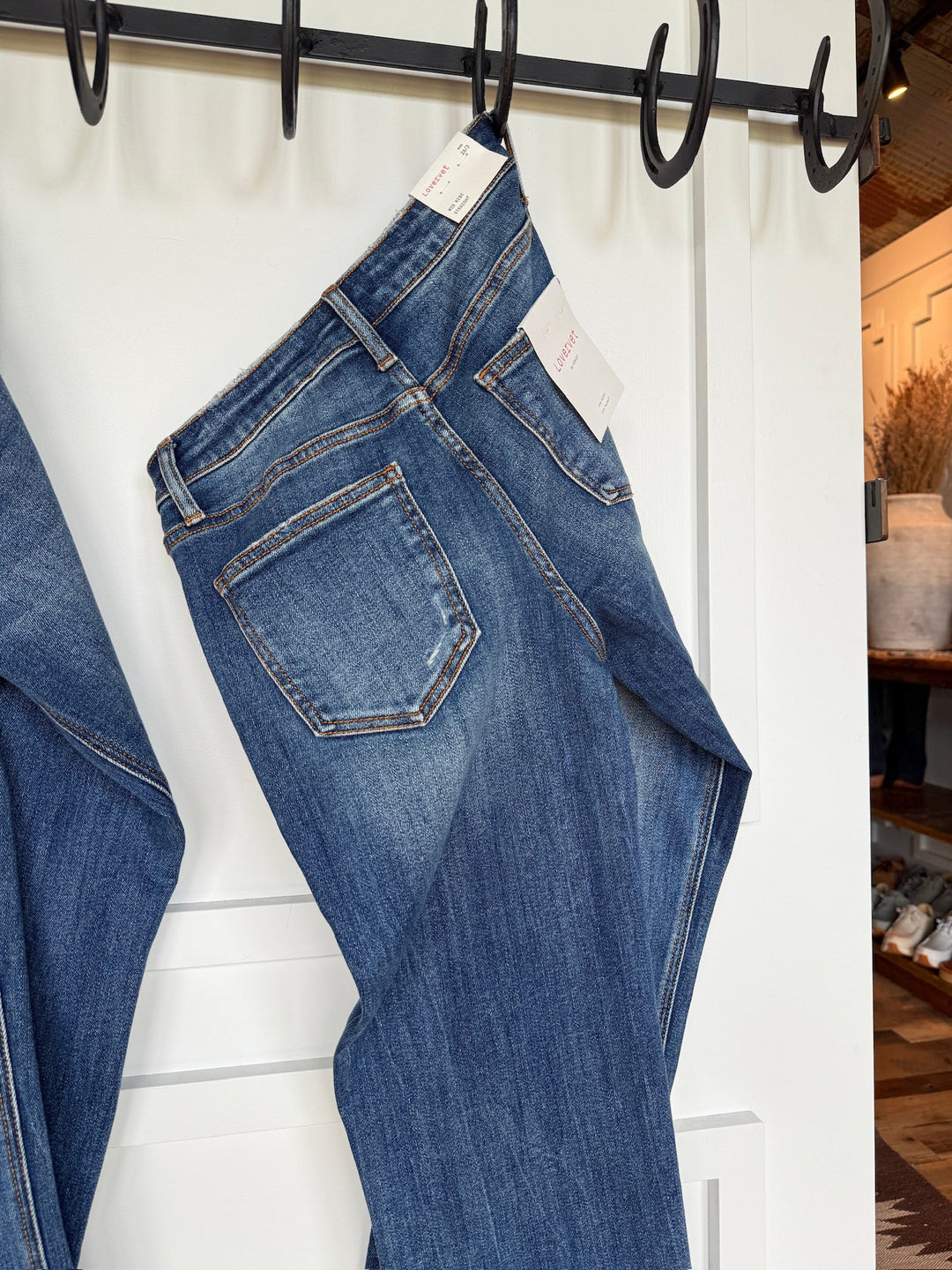 A pair of The Collins Premier Rise Straight Leg Jeans by Savannah Sevens western life{&}style, with tags attached, hangs from a black hook rack on a white door. Another pair in medium dark wash is partly visible to the left.