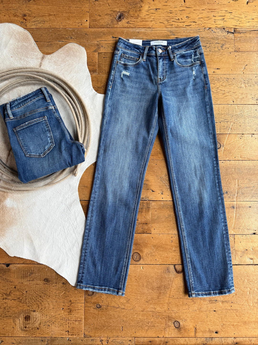 The Collins Premier Rise Straight Leg Jean from Savannah Sevens western life{&}style, shown in a medium dark wash with classic five-pocket design and slight fading, lies flat beside a folded pair, rope, and white animal hide on a wooden floor.