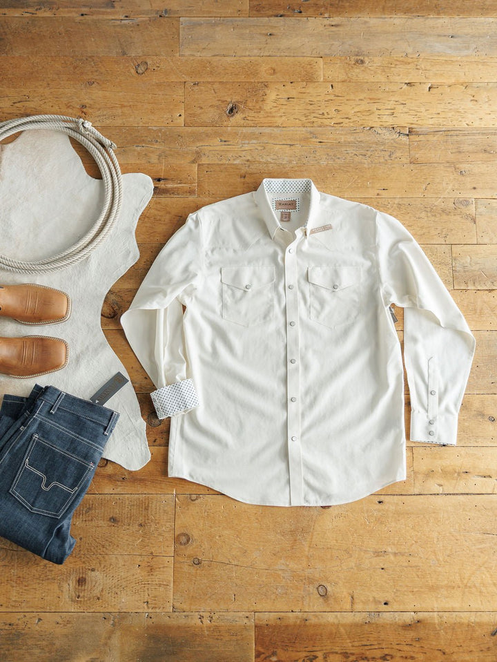 A flat lay features the Collinsville Ariat Men's Natural Pearl Snap by Savannah Sevens western life{&}style, paired with dark blue jeans, a belt, tan shoes, a coiled rope, and a white cowhide on a wooden floor.