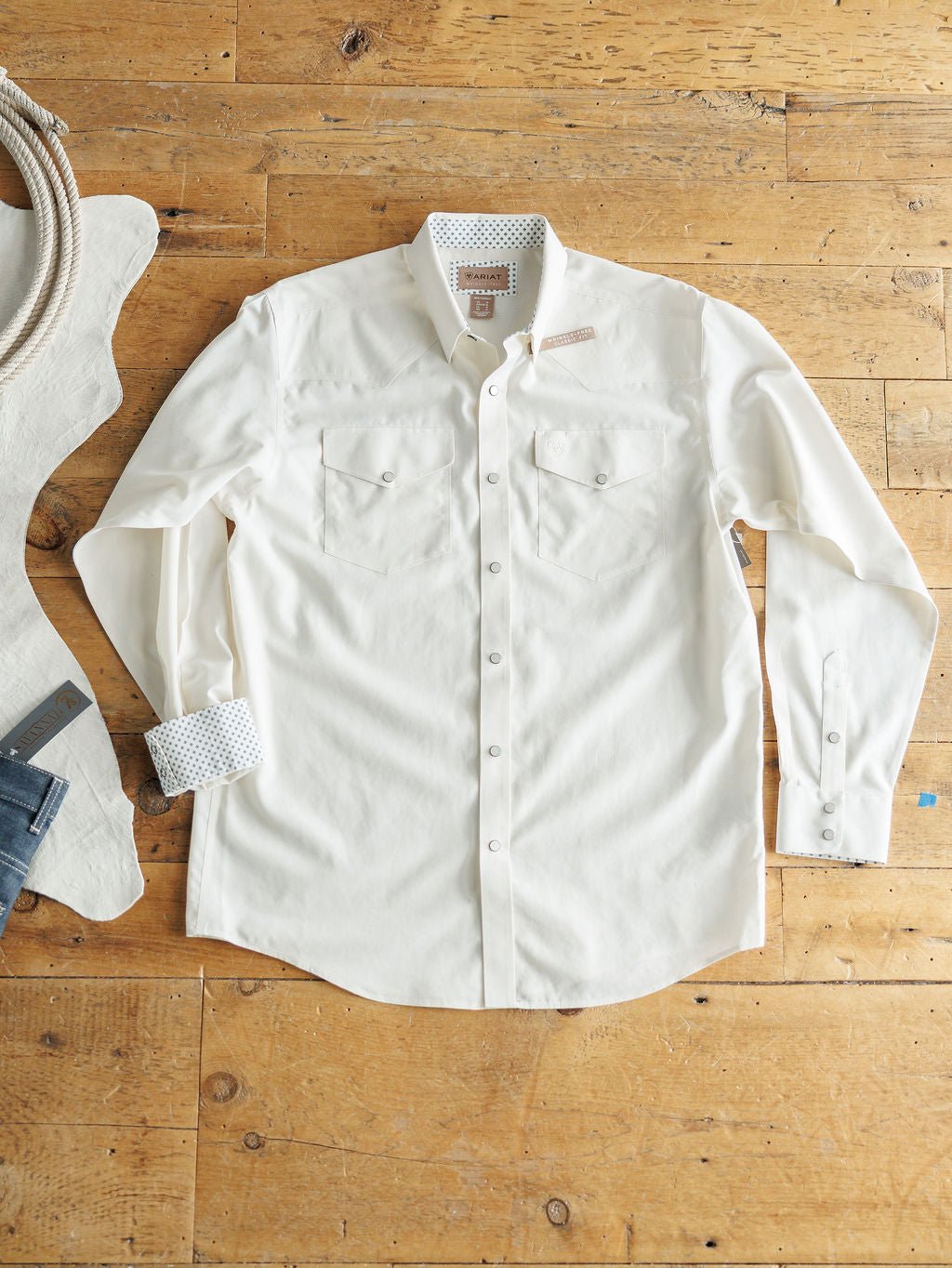 The Collinsville Ariat Men's Natural Pearl Snap shirt by Savannah Sevens western life{&}style, wrinkle-free cotton in off-white, features two chest pockets and patterned inner cuffs, shown flat beside denim jeans and a coiled rope.