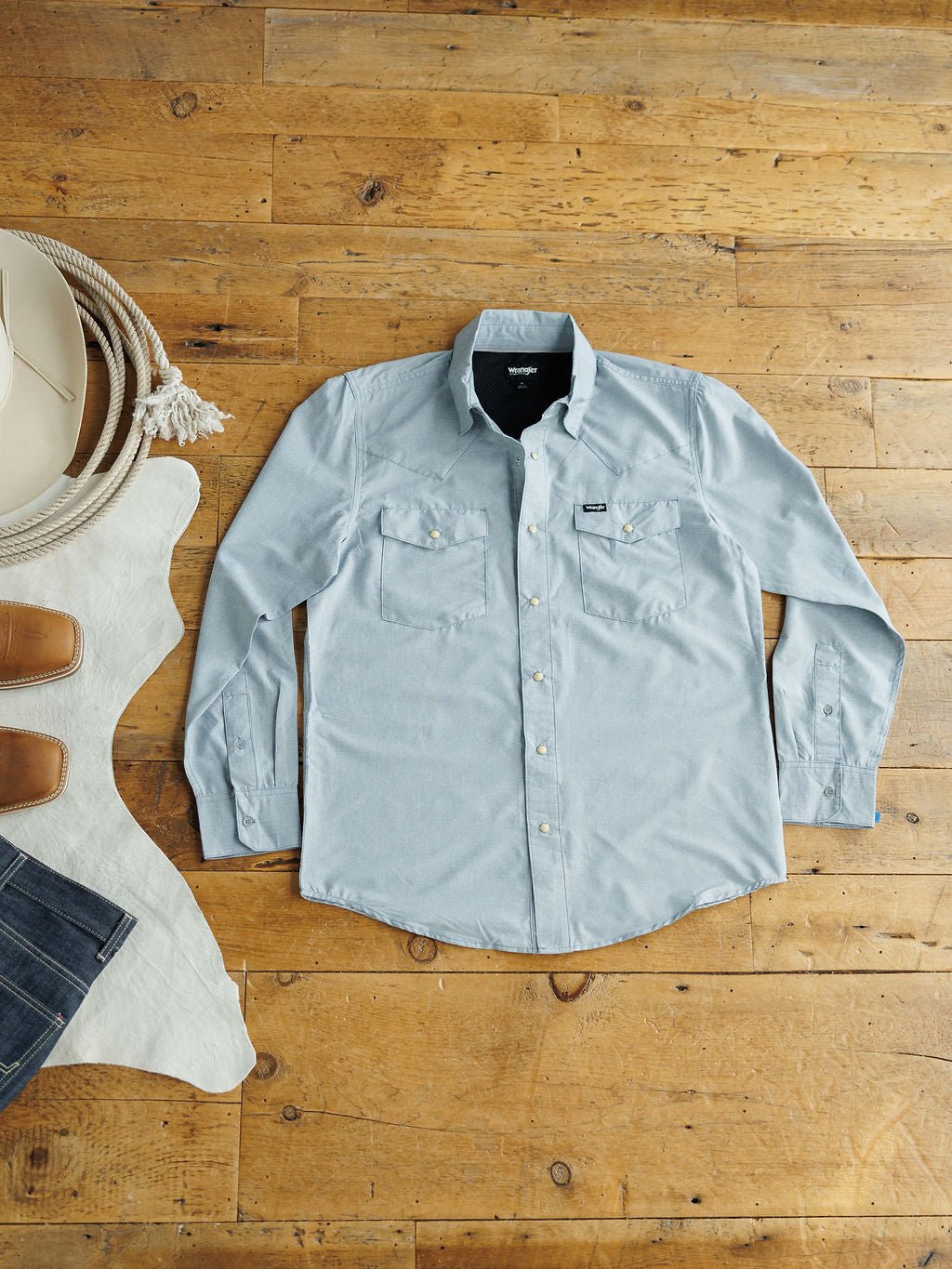 The Colman men's snap button shirt by Savannah Sevens western life&style in light blue is laid flat on a wooden floor beside a white cowboy hat, coiled rope, and a brown and white cowhide rug.