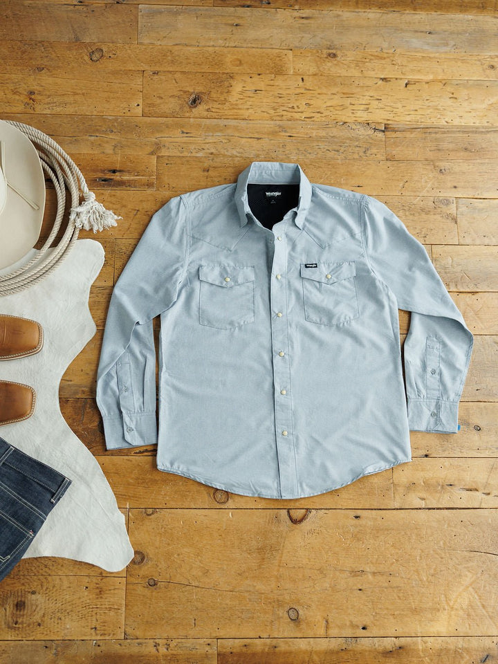 The Colman men's snap button shirt by Savannah Sevens western life&style in light blue is laid flat on a wooden floor beside a white cowboy hat, coiled rope, and a brown and white cowhide rug.