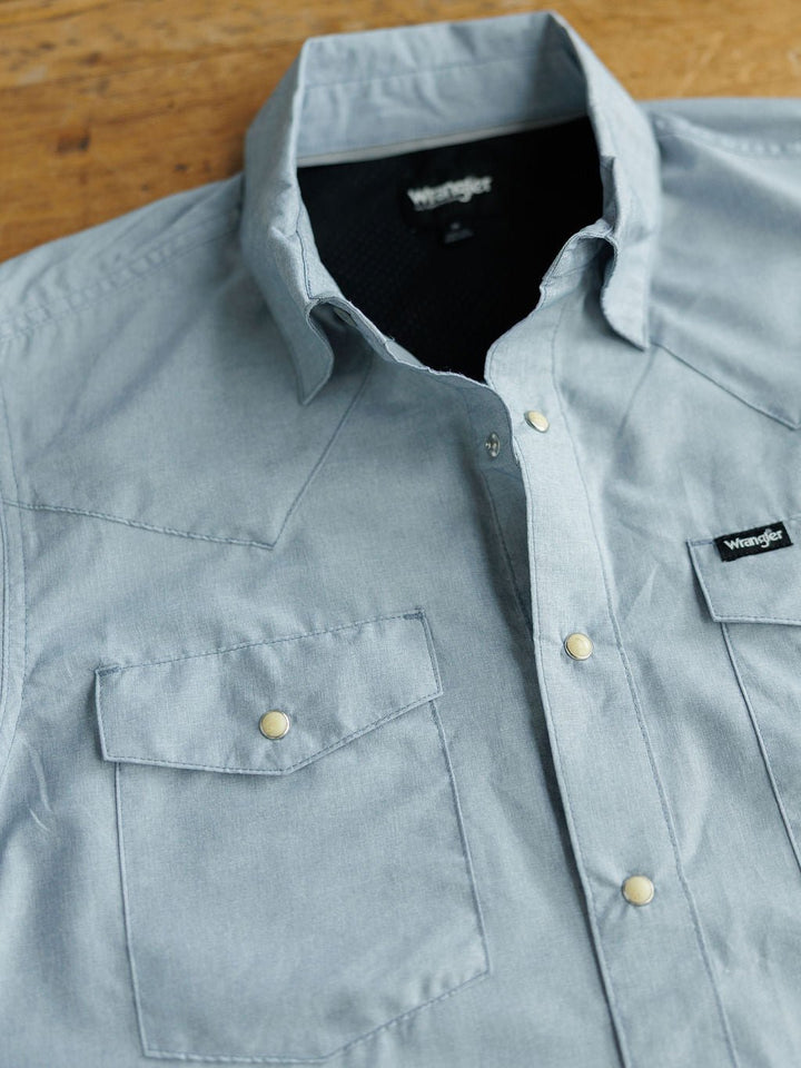A light blue men's snap button shirt, The Colman by Savannah Sevens western life&style, with two chest pockets is laid flat on a wooden surface.