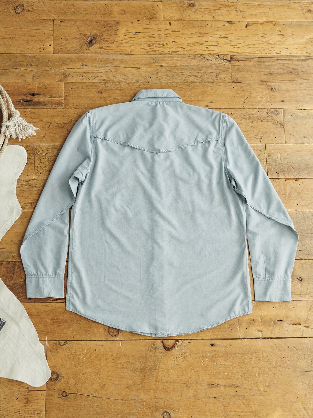 The Colman men's long sleeve shirt by Savannah Sevens western life&style, in light blue moisture-wicking fabric, lies face down on a wooden floor with a back yoke detail, next to beige fabric and a coiled rope on the left.