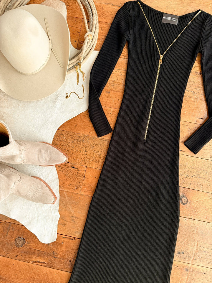 The Colusa Black Ribbed Dress by Savannah Sevens western life{&}style is laid out on a wooden floor next to a beige cowboy hat, white cowhide, and light-colored cowboy boots.