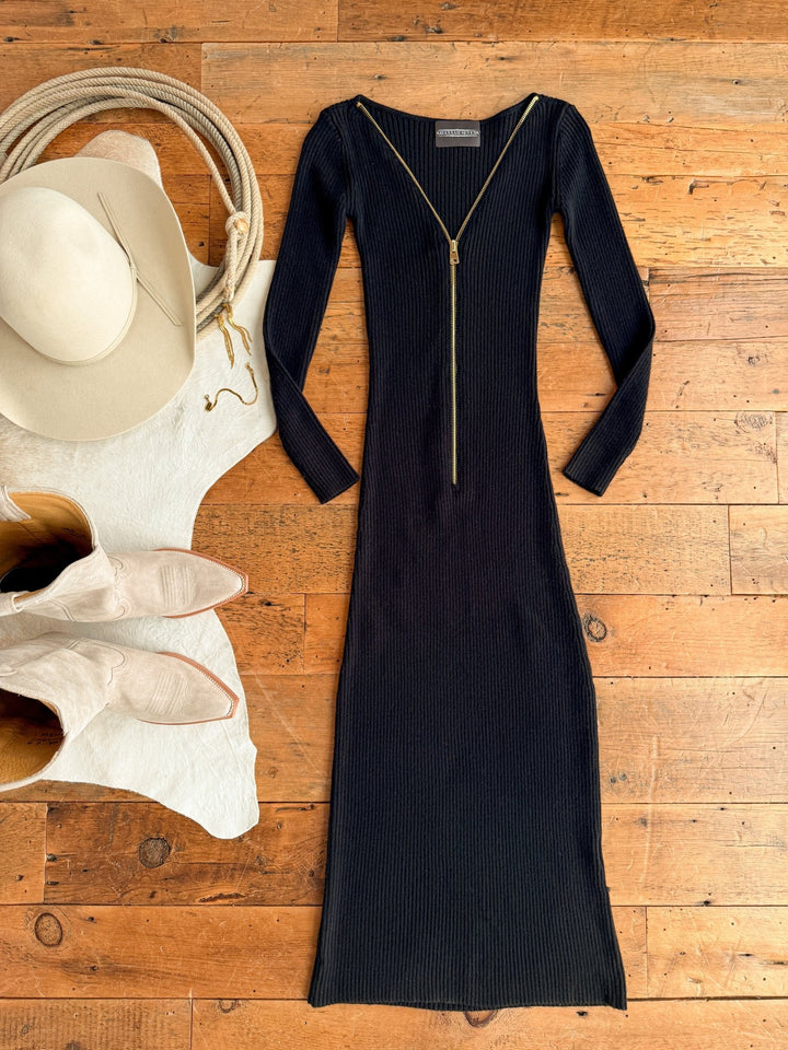 The Colusa Black Ribbed Dress by Savannah Sevens western life{&}style, a long-sleeved bodycon with a front zipper, is laid on a wooden floor beside a cream cowboy hat and light beige boots atop a white cowhide rug.