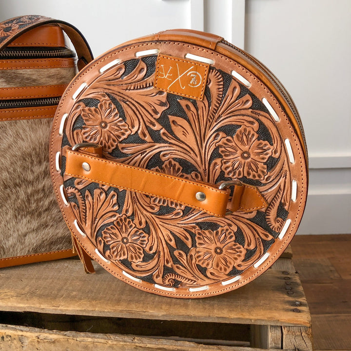 The Colusa Tooled Leather Makeup Case by Savannah Sevens western life{&}style, in tan and dark brown floral tooled leather with a top handle and front strap, rests on wood next to another handmade leather makeup case with unique accents.