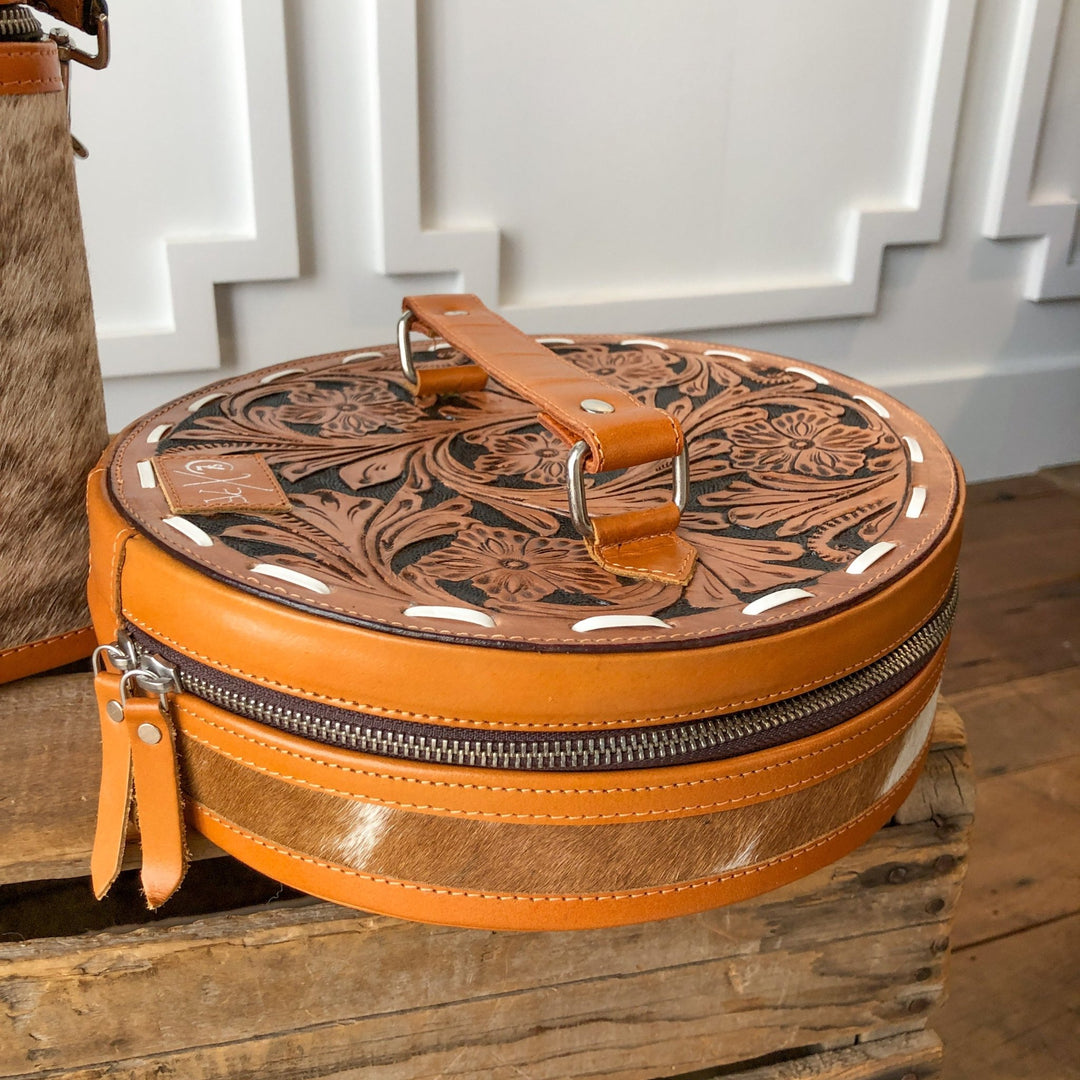 The Colusa Tooled Leather Makeup Case by Savannah Sevens western life{&}style features handcrafted cowhide, a floral tooled lid, brown zipper, and leather handle. Displayed on rustic wood with a white paneled background.