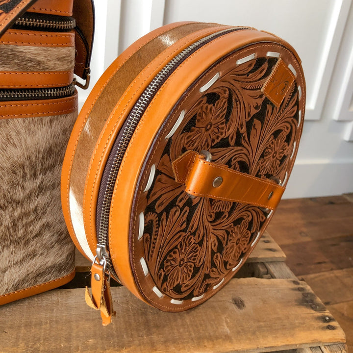 The Colusa Tooled Leather Makeup Case by Savannah Sevens western life{&}style, featuring intricate floral designs, tan accents, a zipper, and a small handle, is shown on wood next to another handmade leather case with cowhide details.