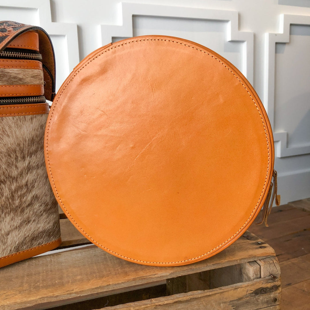 The Colusa Tooled Leather Makeup Case by Savannah Sevens western life{&}style, a round light brown handmade leather bag with zipper, sits on a wooden crate beside a brown and white cowhide-patterned bag with a white trimmed wall in the background.