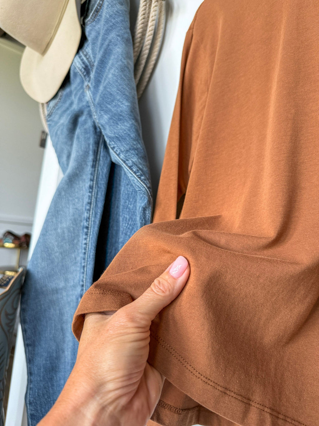 A hand with pink nail polish holds the hem of the Colville Caramel Basic Top by Savannah Sevens western life{&}style. Blue jeans and a beige hat hang on hooks in the background.