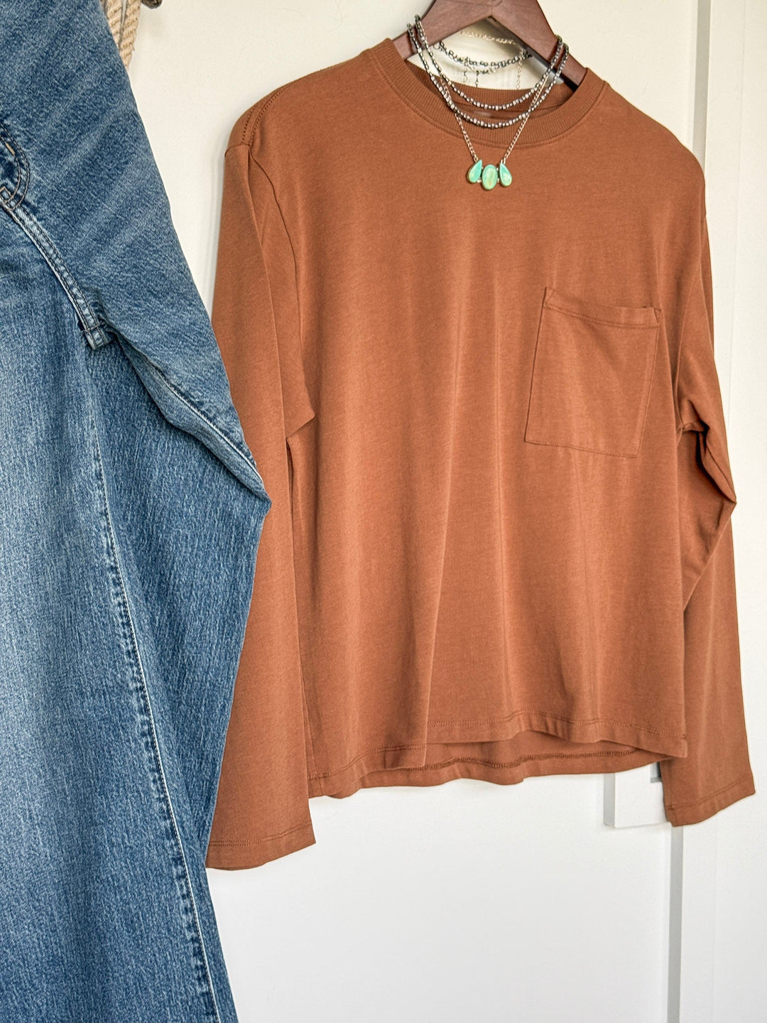 The Colville Caramel Basic Top by Savannah Sevens western life{&}style, featuring long sleeves and a chest pocket in a warm rust shade, hangs beside light blue denim jeans; layered turquoise pendant necklaces are draped over the shirt’s collar.