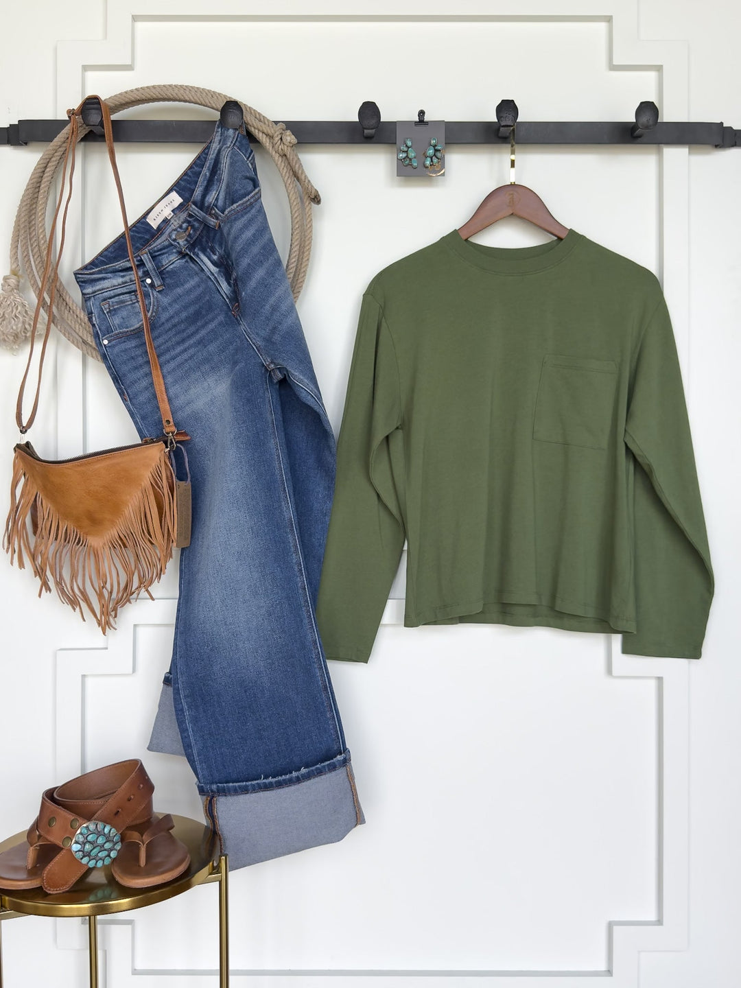 Colville Fern Basic Top by Savannah Sevens western life{&}style in green pima cotton, blue jeans, a brown fringe purse, brown sandals with turquoise detailing, and turquoise earrings are arranged on a white wall and gold stool. The Colville Fern Basic Top features a stretchy feminine fit and a women’s ribbed crew neck for added style.